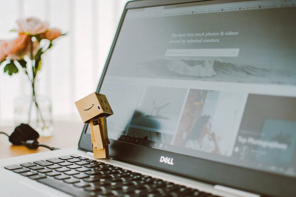 A small toy figure stands on a laptop keyboard with flowers in the background.